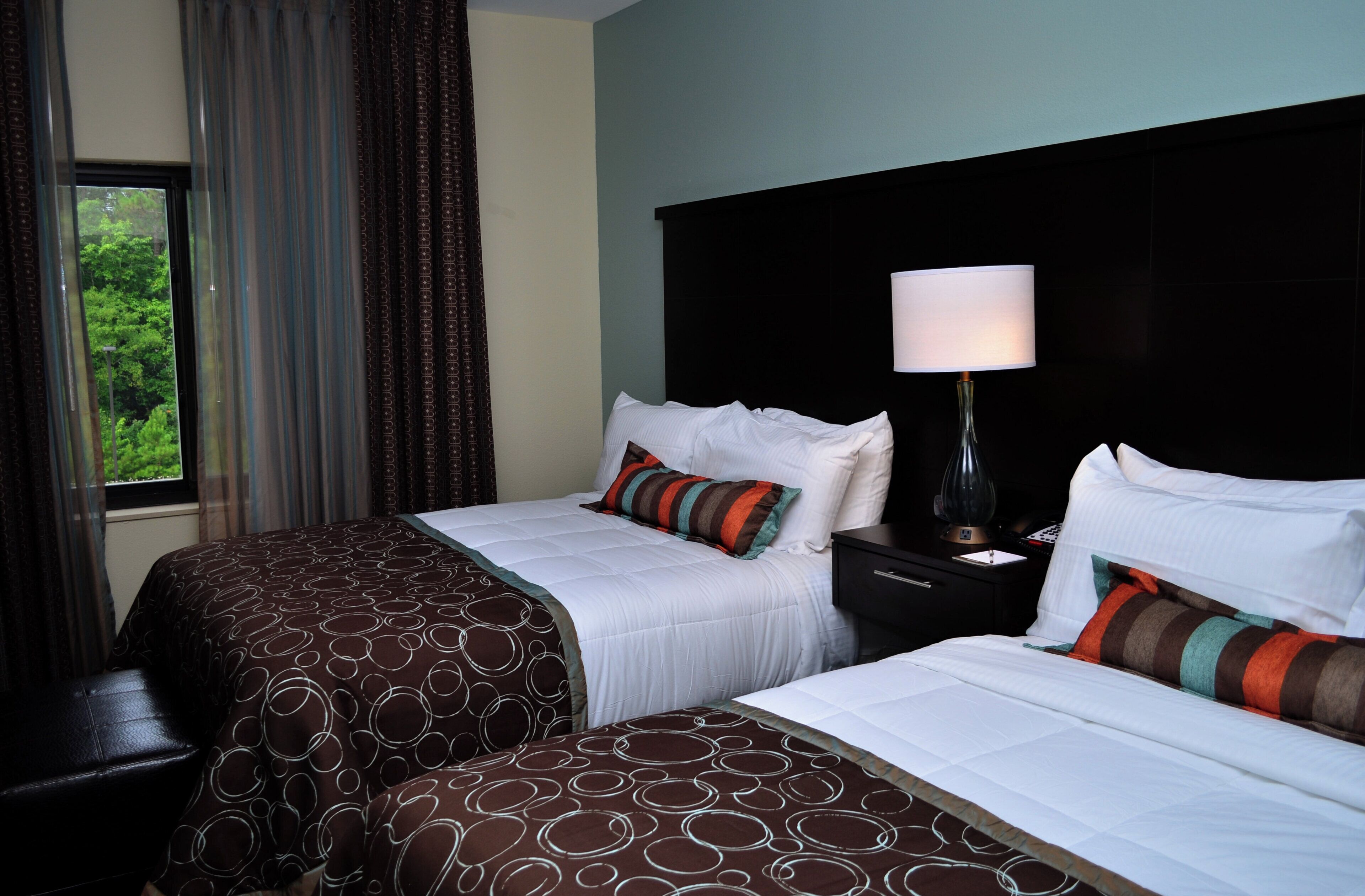 A hotel room with two beds, a nightstand with a lamp, and a window with curtains.