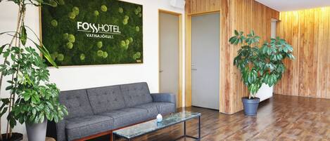 A reception area with a grey sofa, a glass coffee table, and a large green wall with 'FOSSHOTEL' signage.
