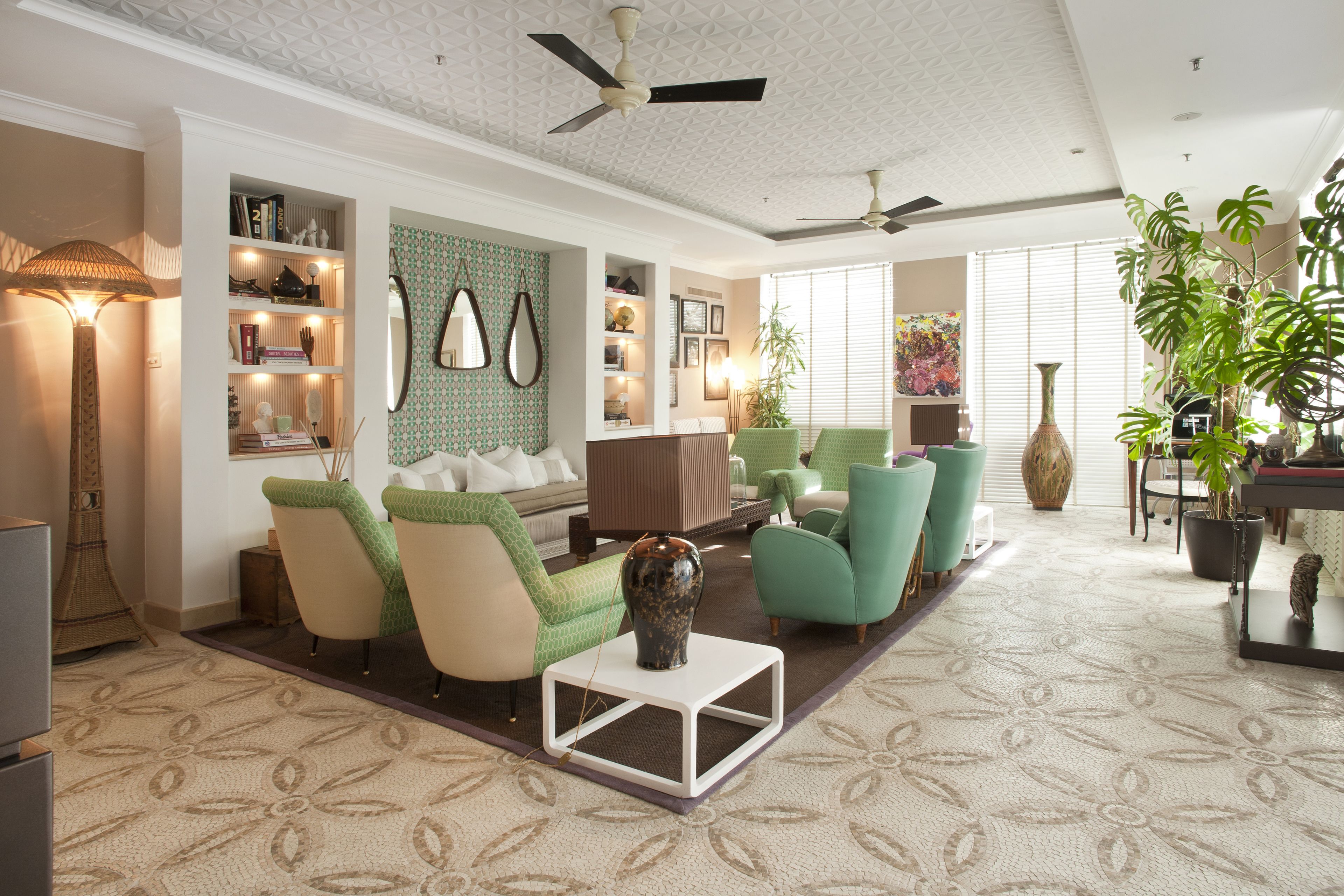 A well-lit living room with a sofa, chairs, a coffee table, and a bookshelf. There are ceiling fans, a large window with blinds, and a potted plant.