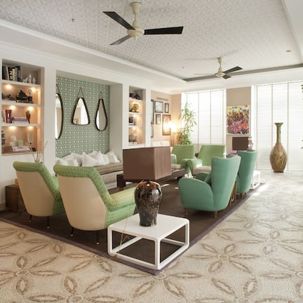 A well-lit living room with a sofa, chairs, a coffee table, and a bookshelf. There are ceiling fans, a large window with blinds, and a potted plant.