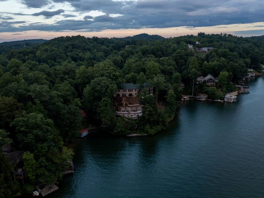 Vista aérea de una propiedad a orillas de un lago, con una casa destacada rodeada de un bosque denso, barcos amarrados y un pequeño muelle.
