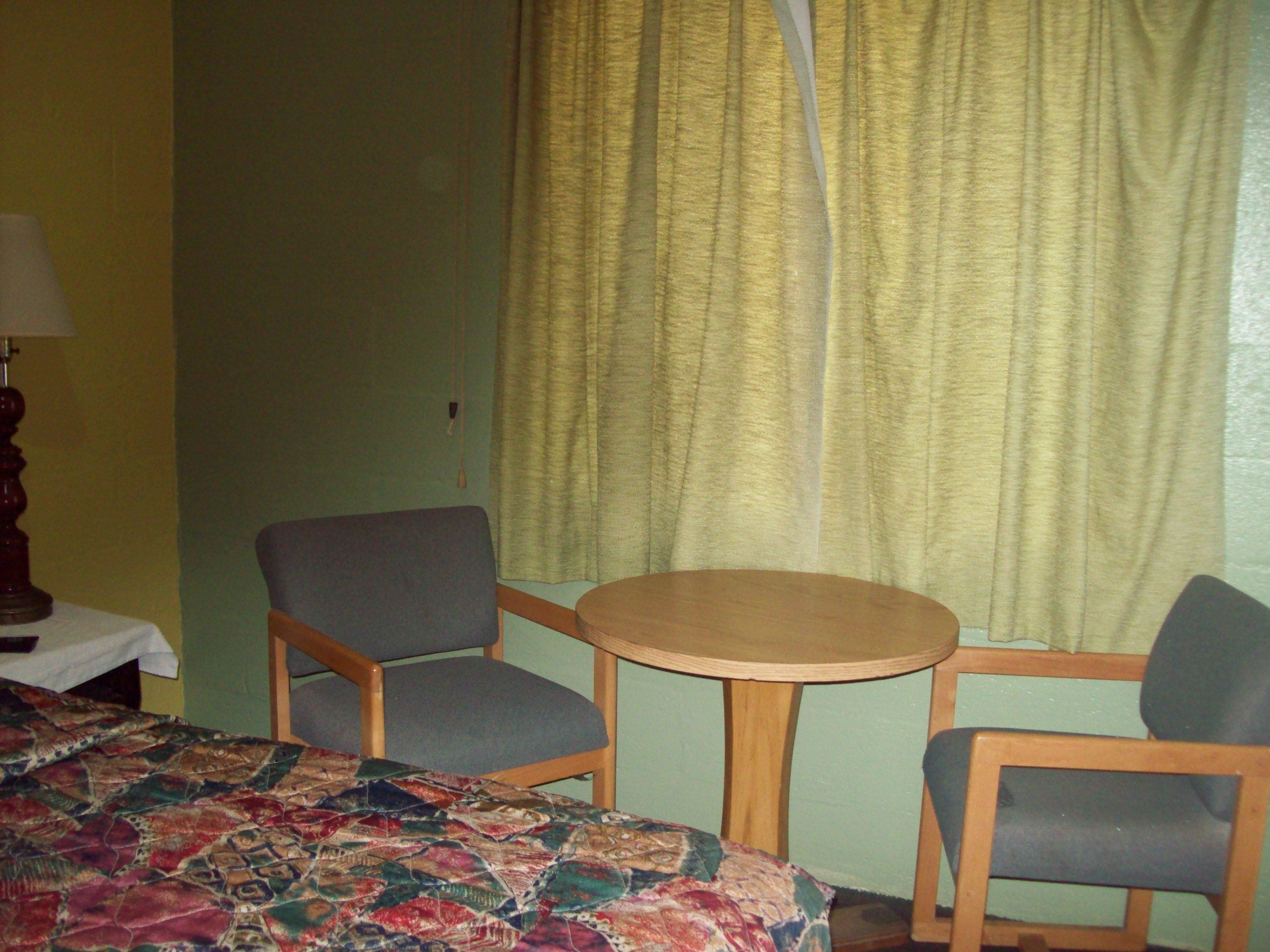 A room with a bed, two chairs, a small table, a lamp, and a curtain.