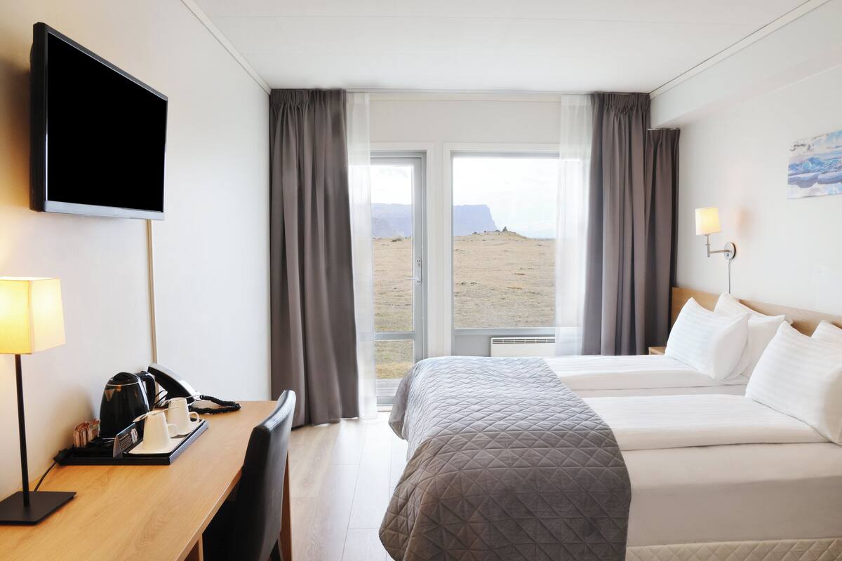 Habitación de hotel con cama, escritorio con teléfono y cafetera, televisor montado en la pared y ventana con cortinas.