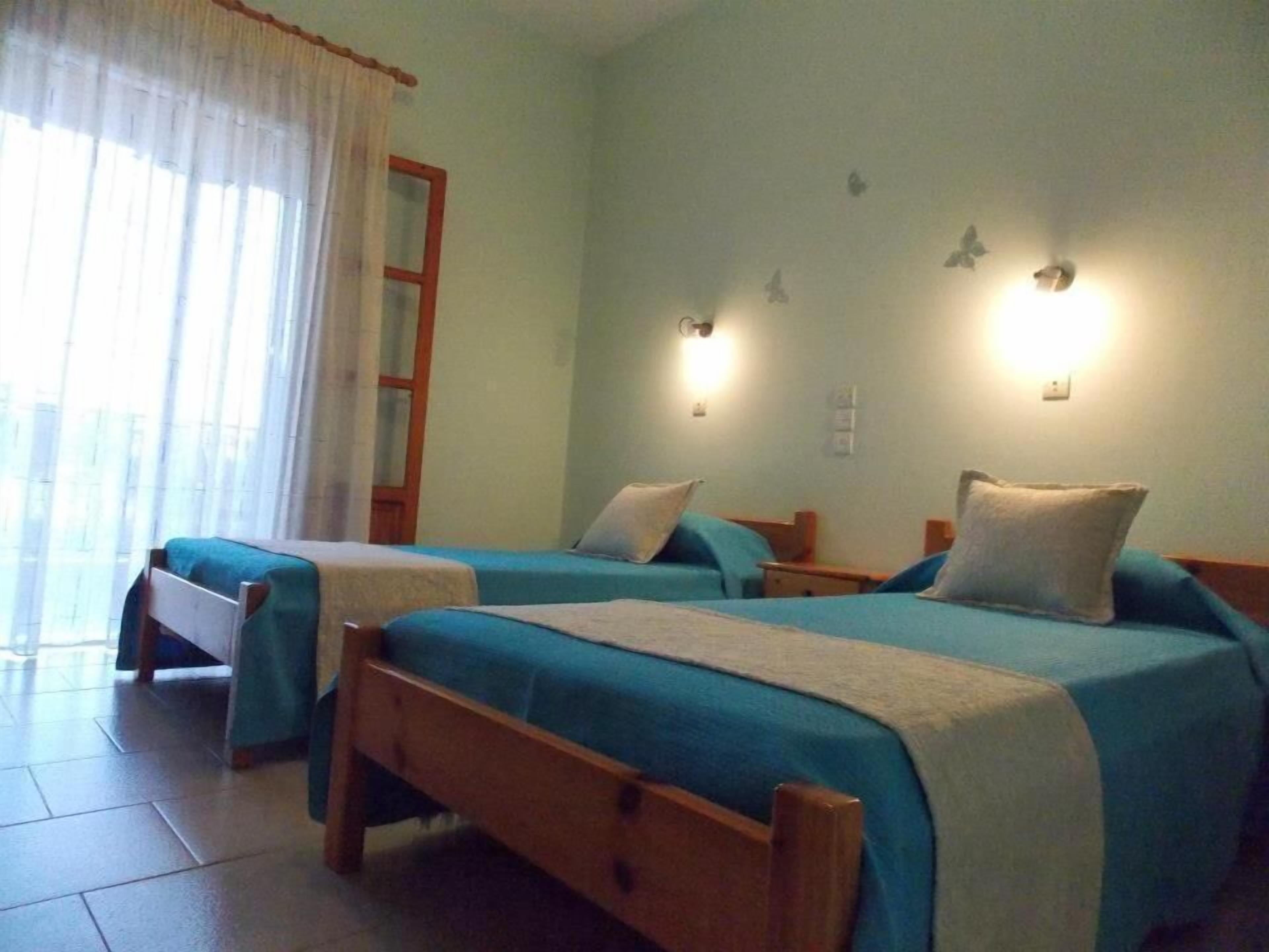 A room with two beds, a wooden floor, a window with curtains, and wall-mounted lights.