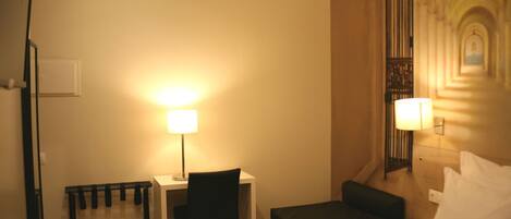 Quarto de hotel com uma cama, uma escrivaninha com cadeira, uma mesinha, ar condicionado e uma luminária fixada na parede.