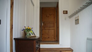 Eine Holztür mit einer kleinen Schrifttafel darüber, eine Holzbank mit einem Blumenstrauß darauf und eine Treppe, die zu einer Zwischenebene führt.