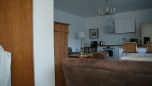 Ein Wohnzimmer mit braunem Sofa, einem Holzschrank, einem Esstisch mit Stühlen, einer Küchenzeile mit Schränken, einem Herd und einer Kristallleuchter.