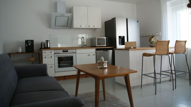 Eine moderne Küche mit weißen Schränken, einer Mikrowelle, einem Backofen und einem Kühlschrank. Ein Holztisch mit einem Einmachglas steht daneben, und zwei Korbstühle ergänzen das Bild.