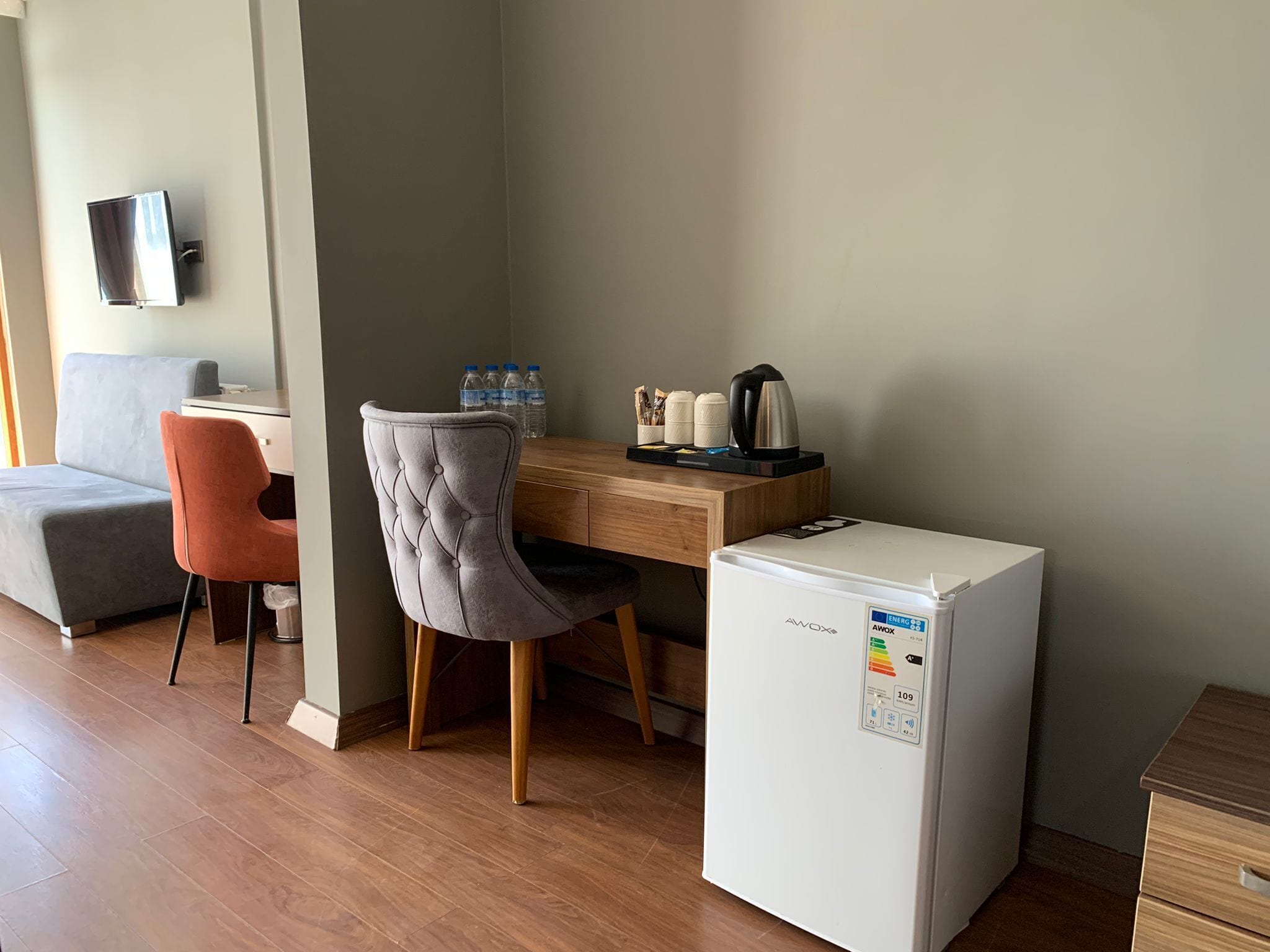 A compact hotel room with a wooden desk, a grey upholstered chair, a white refrigerator, and a grey sofa.