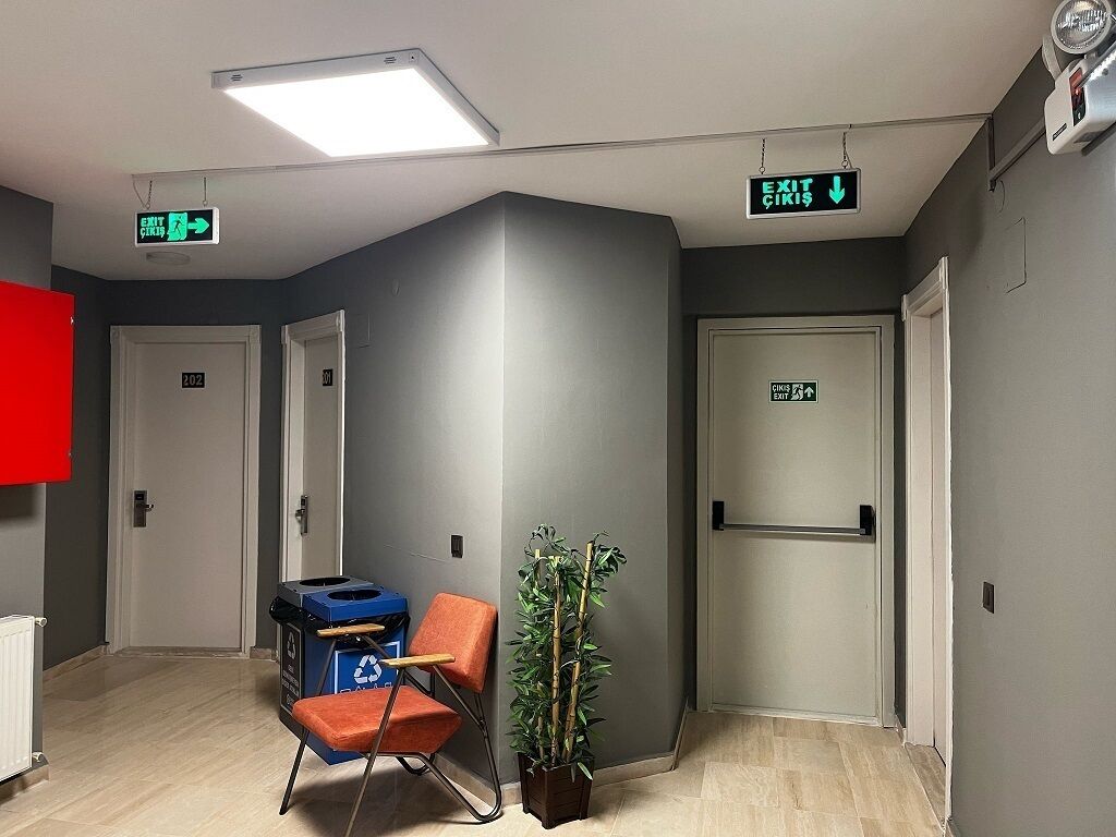 Un pasillo con paredes grises, un armario rojo y una planta. Hay puertas con los números 202 y 301, y una señal de salida.
