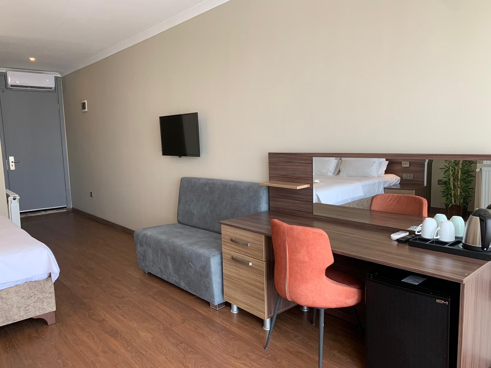 Une chambre d’hôtel comprenant un lit, un canapé gris, un bureau en bois avec une chaise, une télévision et un petit réfrigérateur.