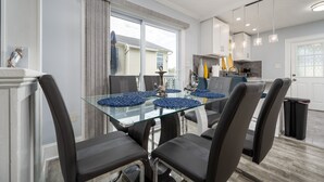 A modern dining area with a glass table and black chairs, a kitchen in the background, and a view of a house through large windows. - Parrot Bay 2 bedroom Condo in Downtown OC with pool near the beach (Ocean City)
