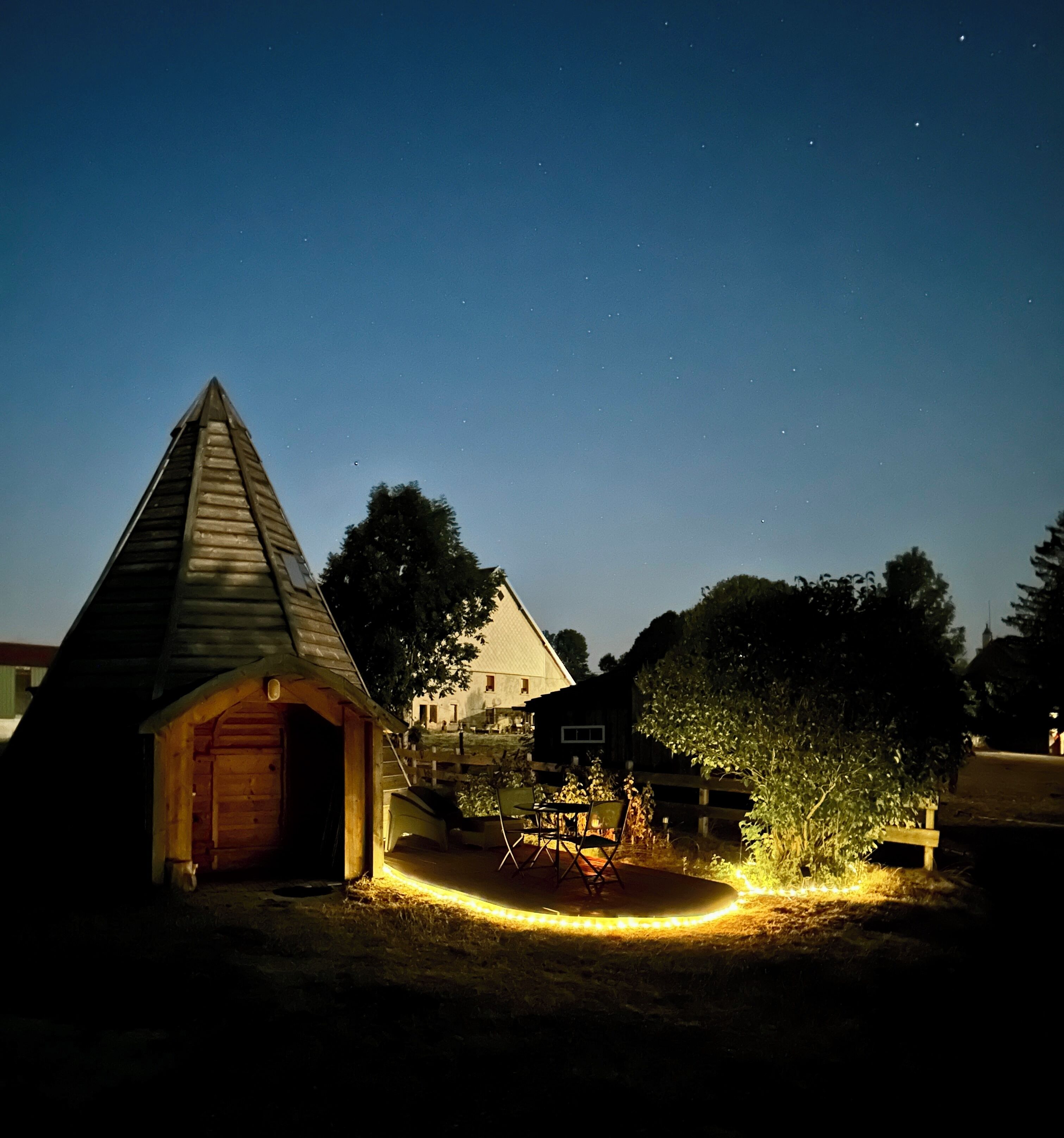 Una cabaña de madera con techo inclinado, un patio cercado y una pequeña mesa con sillas.