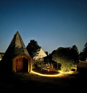 A wooden cabin with a sloped roof, a fenced yard, and a small table with chairs.