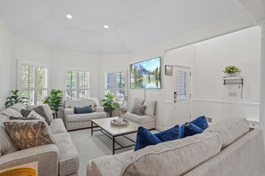 Ein geräumiges Wohnzimmer mit einem großen Flachbildfernseher an der Wand, einer Sofagarnitur, einem Couchtisch und Blick auf einen Bergsee durch große Fenster.
