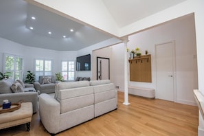 A spacious living room with a large grey sofa, a smaller armchair, and a wooden bench. There are potted plants and a flat-screen TV mounted on the wall. - 2 short blocks  to the beach. FULLY RENOVATED HOUSE  WITH NEW HOT TUB. (Point Pleasant Beach)