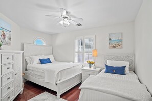 A bedroom with two beds, a ceiling fan, a window with blinds, and artwork on the walls.