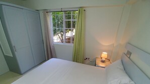 A bedroom with a bed, a nightstand with a lamp, a wardrobe, and a window with curtains. - 1 Modern A/C apartment with pool. Walking distance from beach, shop & restaurant (Castries)