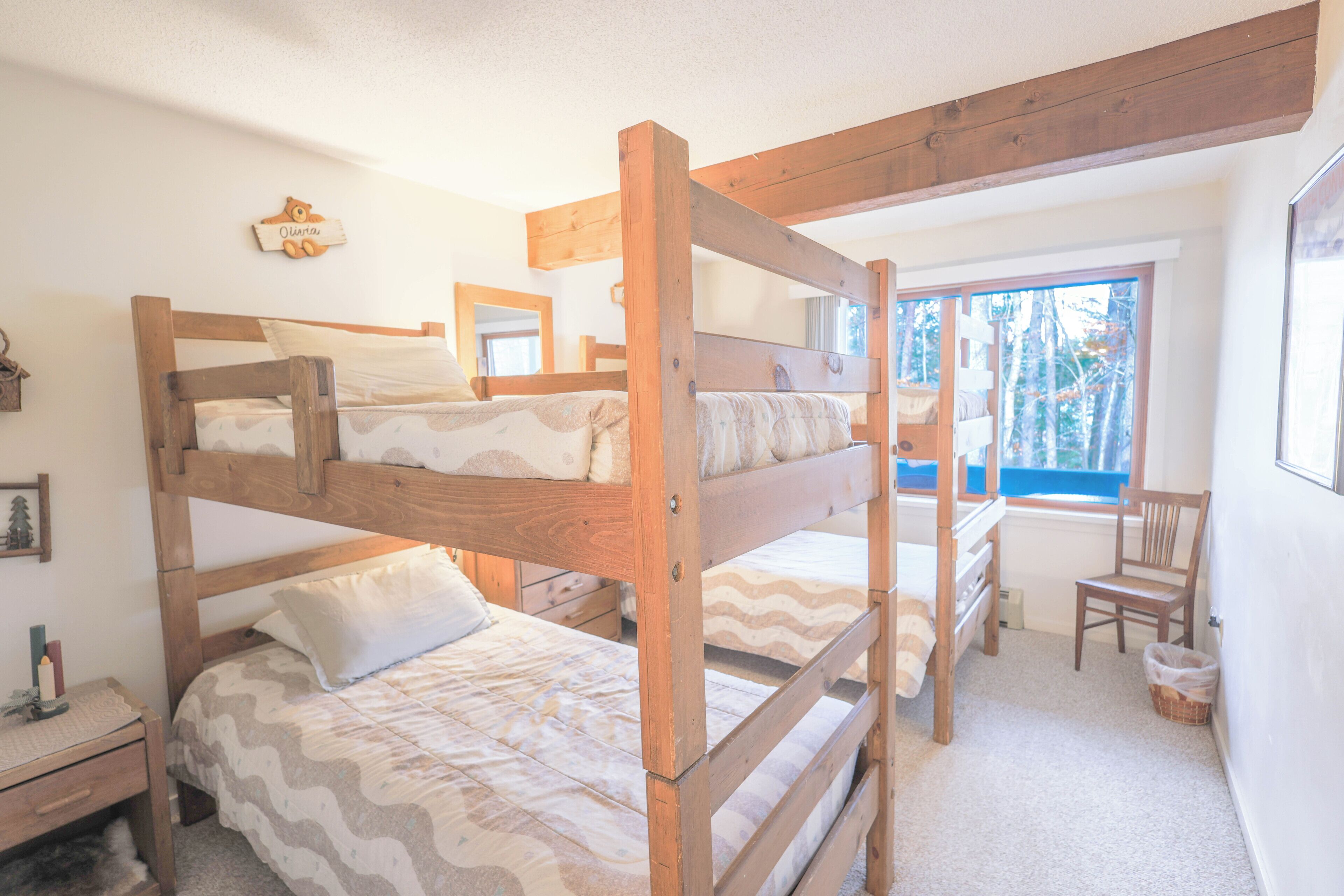 Dormitorio con literas, cabecera de madera, mesita de noche pequeña, silla y ventana con vista a árboles.