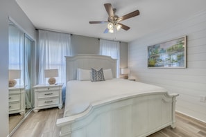 Un dormitorio con una cama blanca, un ventilador de techo, una mesita de noche con lámpara y un cuadro en la pared.