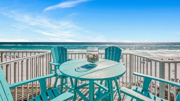 A turquoise deck with a round table and chairs, overlooking a beach with the ocean in the background.