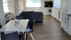 A modern kitchen with a dining area, a grey sofa, and a TV mounted on the wall.