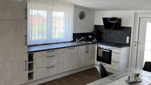 A modern kitchen with grey cabinets, a black countertop, a window with sheer curtains, a wall clock, and a dining area with a table and chairs.