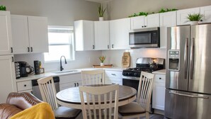 Una cocina moderna con armarios blancos, electrodomésticos de acero inoxidable, una mesa de comedor con sillas y una manta amarilla en un sofá.