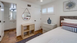 Ein Schlafzimmer mit Bett, Nachttischen, einem Schminktisch, einem Spiegel, einem Fenster mit Jalousien und einem Deckenventilator.