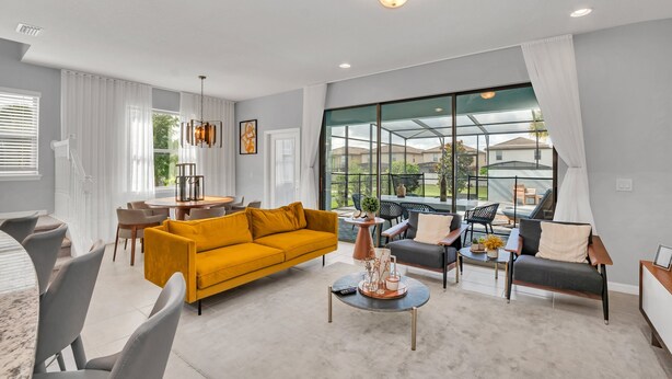 A modern living room with a yellow sofa, grey armchairs, and a coffee table. There is a dining area with a table and chairs, and a large window with a view of the outdoors. - EV_8900 - 5BR / 4.5BA Villa w/ Private Pool, Game Room & Resort Access (Kissimmee)