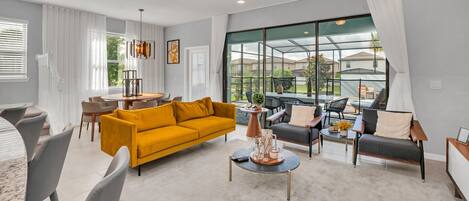 Ein modernes Wohnzimmer mit einem gelben Sofa, grauen Sesseln und einem Couchtisch. Es gibt eine Essecke mit Tisch und Stühlen sowie ein großes Fenster mit Blick ins Freie.