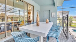 A modern outdoor dining area with a white table and blue chairs, large windows, and a view of the neighborhood. - EV_8900 - 5BR / 4.5BA Villa w/ Private Pool, Game Room & Resort Access (Kissimmee)