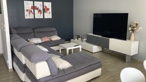 Een moderne woonkamer met een grijze bank, een witte salontafel en een flatscreen televisie op een witte standaard.