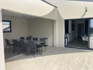 A modern outdoor dining area with a glass table and chairs, a potted plant, and a view into the adjacent room.