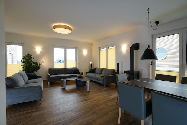Ein modernes Wohnzimmer mit einer Sofagarnitur, einem Esstisch und einem großen Fenster mit Jalousien.