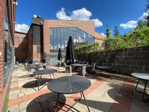 An outdoor patio with wicker furniture and black umbrellas, a brick building with large windows, and a clear blue sky. - TH City Suite 3 (Tampere)