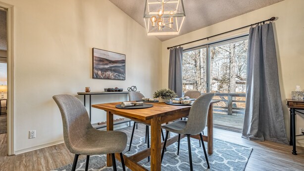 A dining area with a wooden table, four chairs, a chandelier, a painting, and a view through large windows. - Golf Lover's Getaway! Cozy 2BR/2BA Townhome, Walk to DeSoto Golf Club! (Hot Springs)