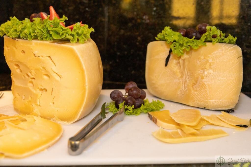 Dos ruedas grandes de queso con lechuga y uvas encima, acompañadas de virutas de queso y un cuchillo sobre un plato blanco.