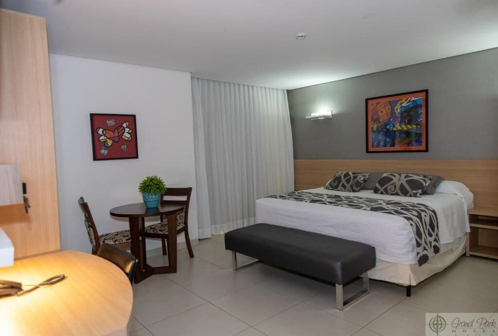 A hotel room with a bed, a small table with a chair, a bench, and two paintings on the walls.