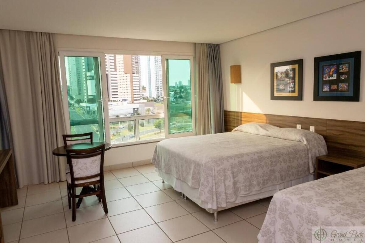 Ein Hotelzimmer mit zwei Betten, einem Esstisch mit Stuhl, großen Fenstern mit Vorhängen und gerahmten Bildern an der Wand.