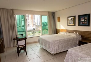 Una habitación de hotel con dos camas, una mesa de comedor con una silla, ventanas grandes con cortinas y cuadros en la pared.