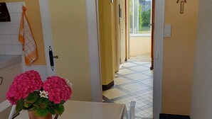 Un pasillo con una puerta blanca, una planta en maceta con flores rosadas y un reloj en la pared.