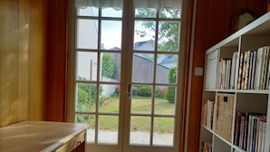 Una habitación con un escritorio de madera, una estantería y un ventanal con vistas a un jardín y casas.