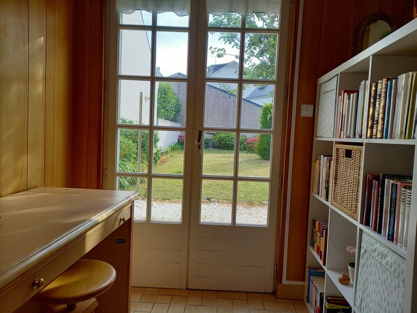 Een kamer met een houten bureau, een boekenkast en een groot raam met uitzicht op een tuin en huizen.