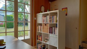 Una habitación con un ventanal grande, una estantería llena de libros, una mesita con una taza y un cuaderno, y una puerta que da a una escalera.