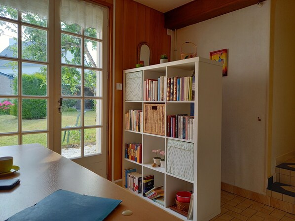 Een kamer met een groot raam, een boekenplank vol boeken, een klein tafeltje met een kop en een notitieboek, en een deur die leidt naar een trap.