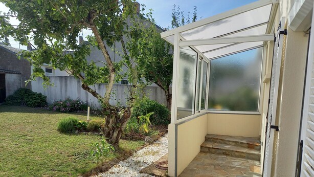 Un invernadero con un sendero de piedra que lleva a una puerta, rodeado de un jardín con un árbol y diversas plantas.