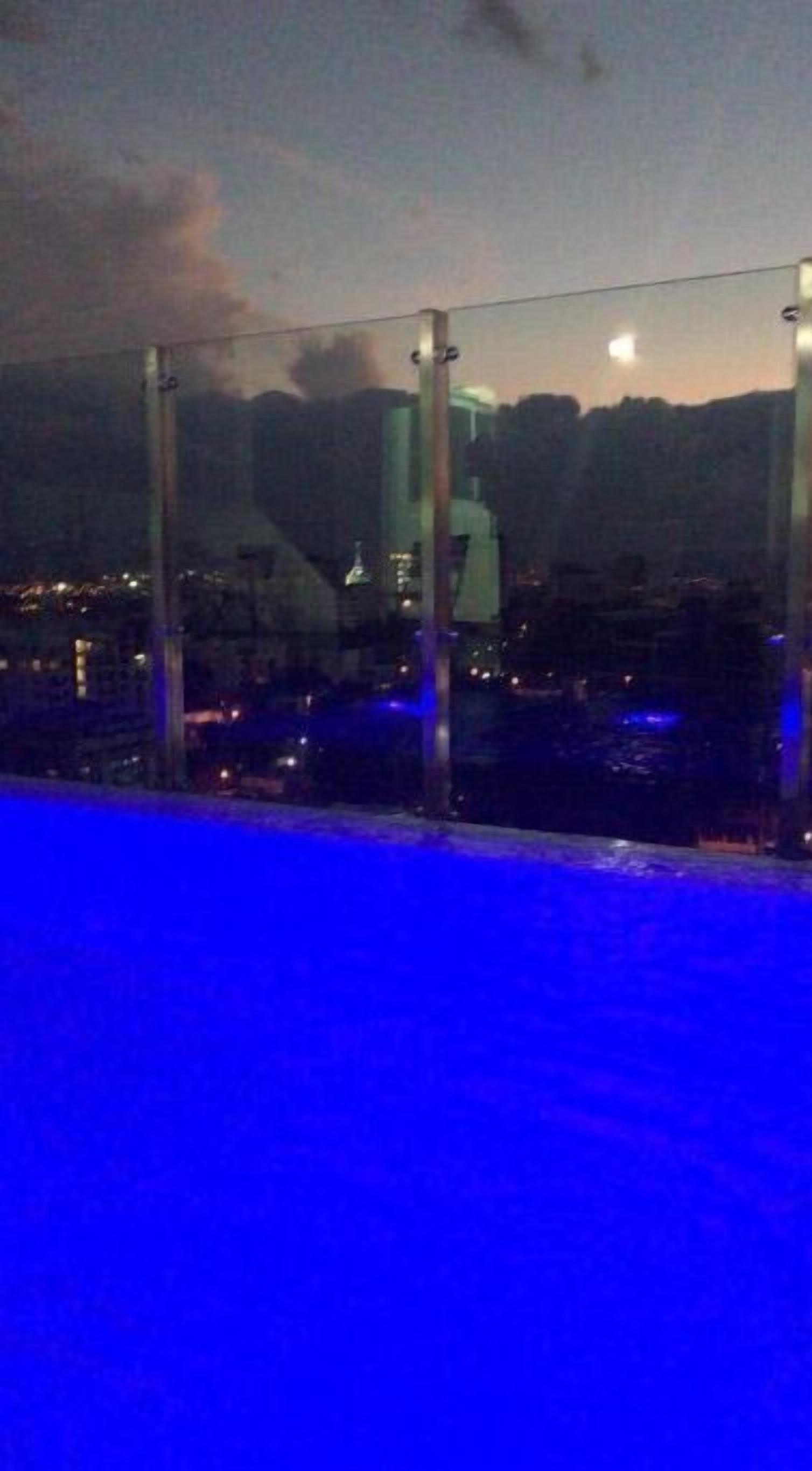 Vue nocturne depuis un balcon donnant sur une piscine, avec en contrebas un paysage urbain composé d’immeubles et, au loin, une chaîne de montagnes.
