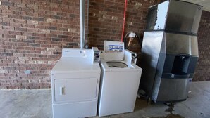 A laundry area with a Whirlpool washer and dryer, a stacked refrigerator, and a brick wall. - Scottish Inns Jonesboro AR  (Jonesboro)