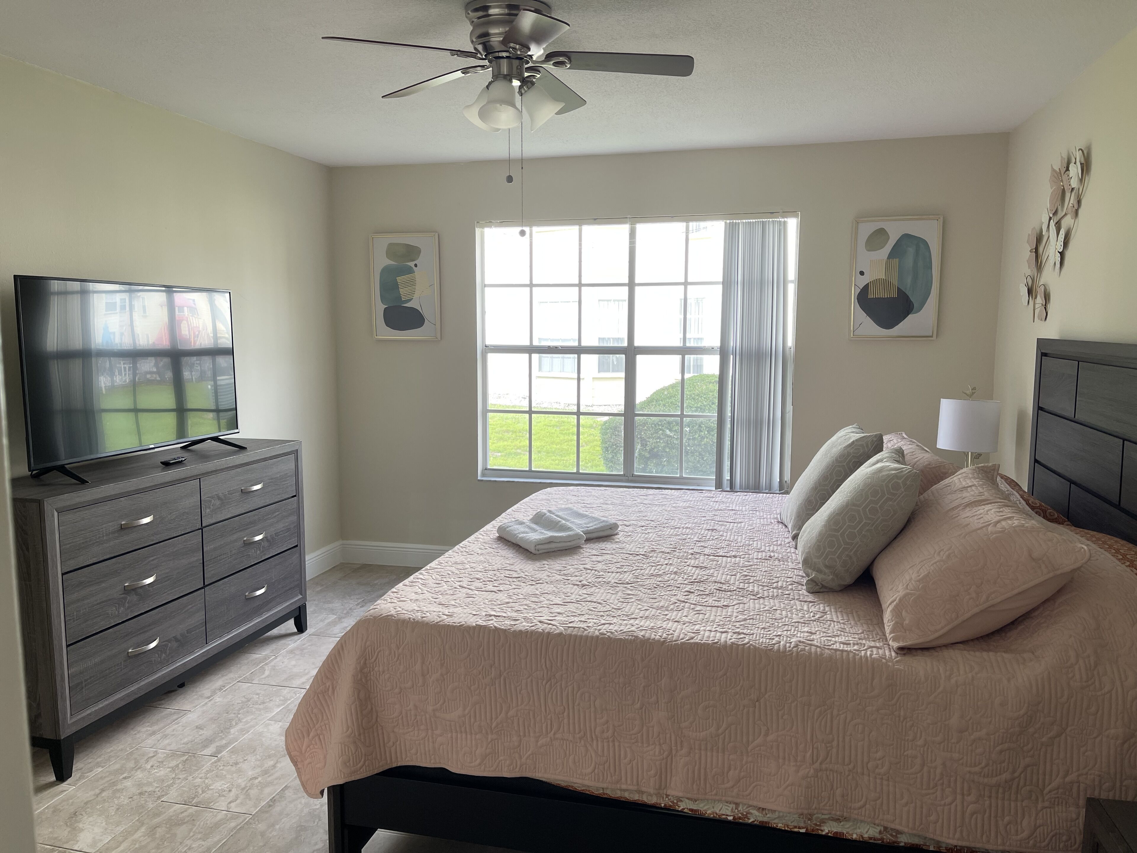 A bedroom with a bed, a dresser, a television, and a window with a view of greenery.